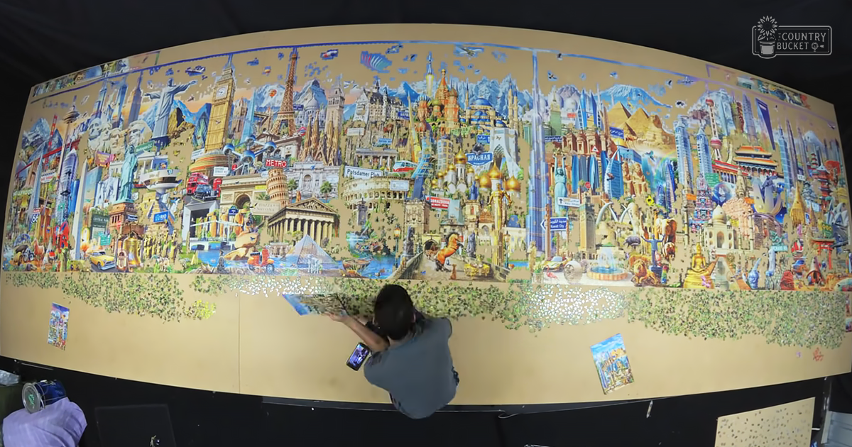 An indoor scene with an adult male assembling a large, colorful world landmarks puzzle under artificial lighting.