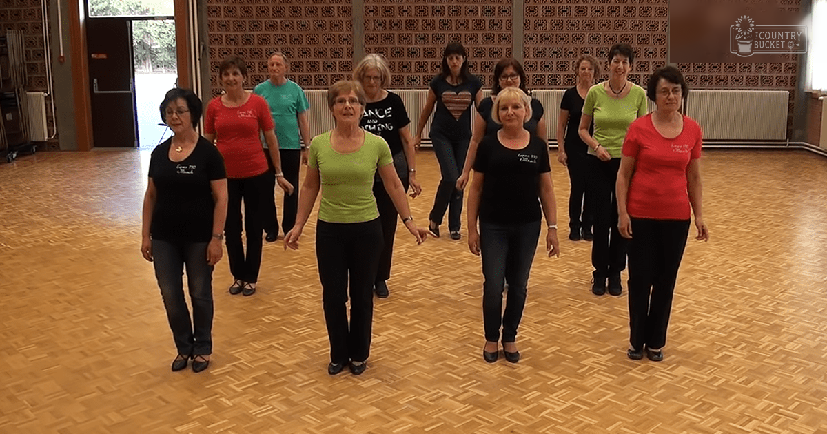 Group of adult females dancing indoors in a dance studio with wooden flooring and patterned wallpaper, natural light from windows, spring or summer setting.