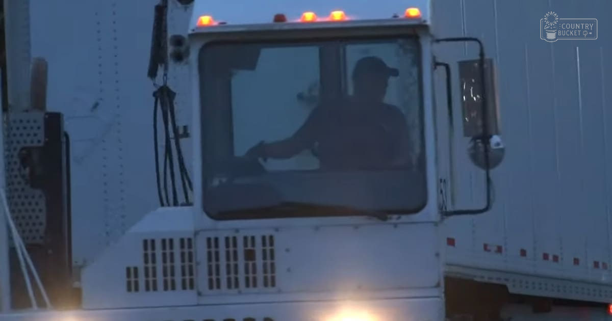 An adult male in a dark baseball cap, light-colored t-shirt, seated inside a white semi-truck cab at night with artificial lighting and orange warning lights visible.