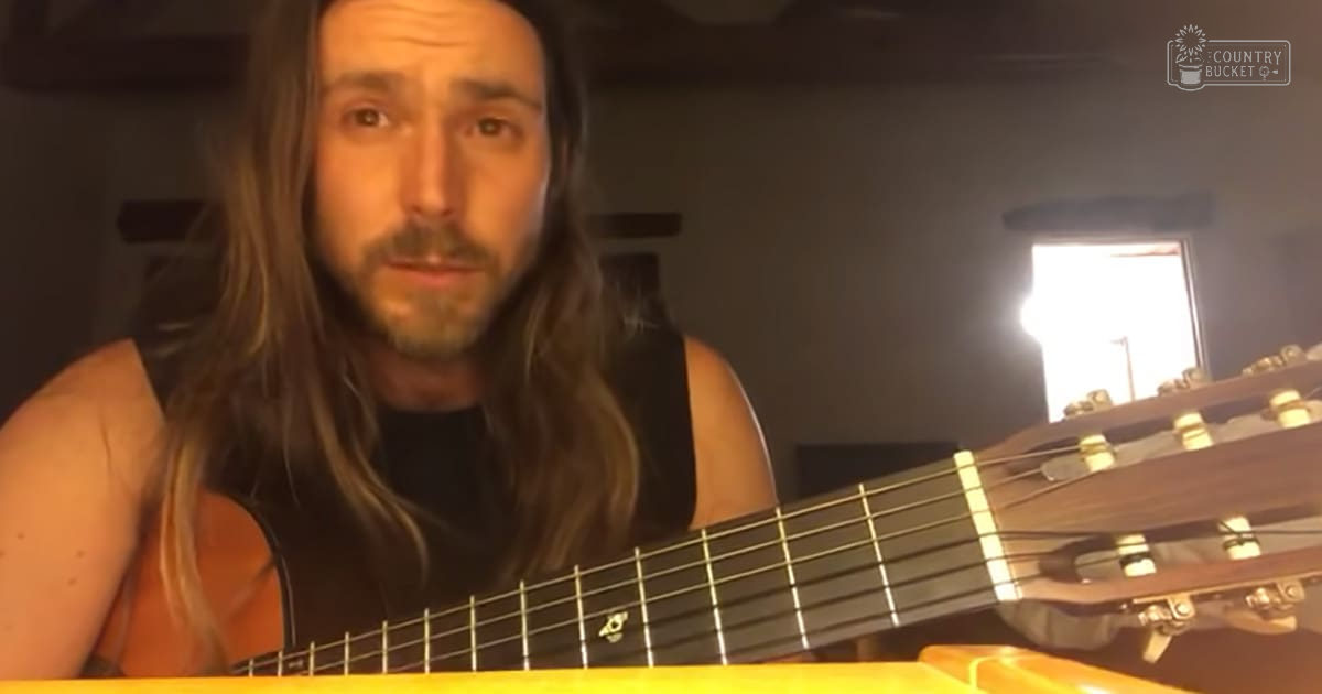 An adult male playing an acoustic guitar in a home setting, wearing a black sleeveless top with long dark brown hair. The background suggests daytime indoor lighting with natural light streaming through a window.
