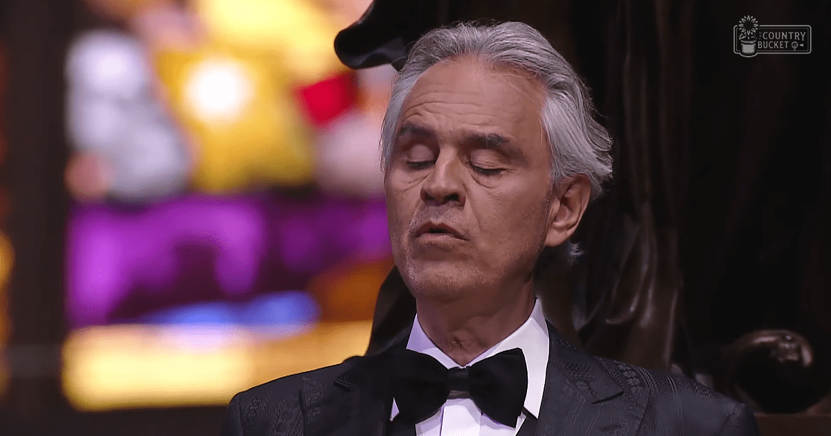 An older adult male wearing a black tuxedo with intricate embroidery stands against a stage setting with colorful lighting.