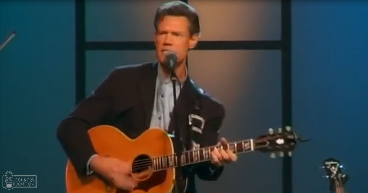 A middle-aged male singer performing on stage with an acoustic guitar and microphone at an indoor venue.