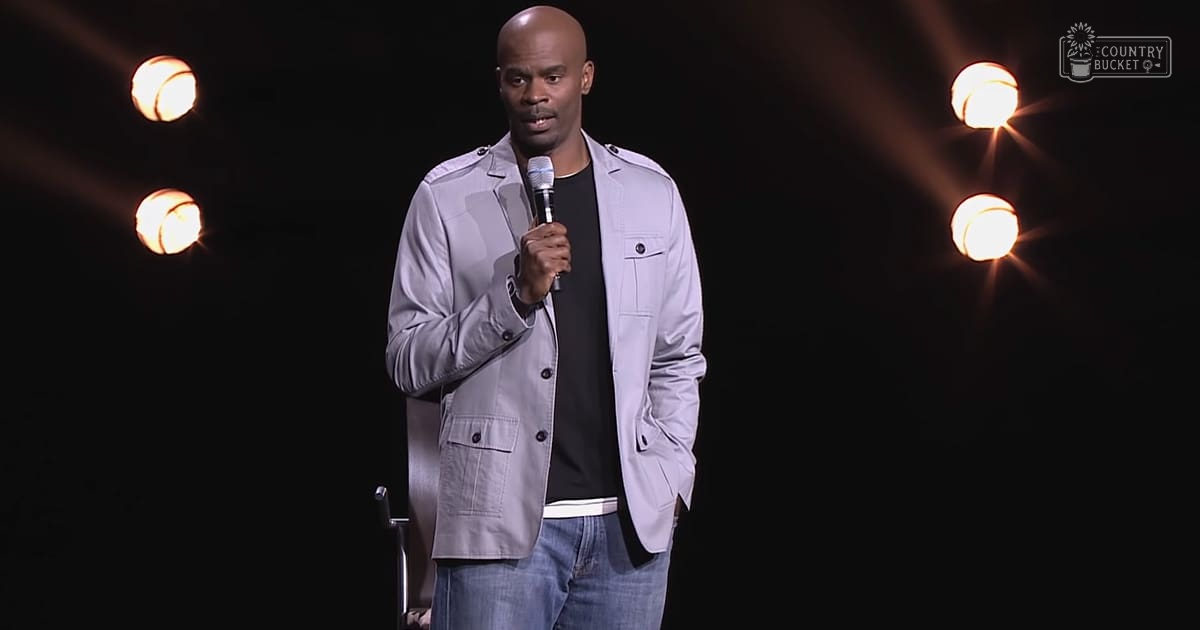 A middle-aged male comedian performing stand-up comedy in a dimly lit indoor setting with spotlights, wearing a light gray jacket over a black shirt and blue jeans, holding a microphone.