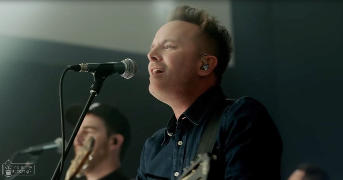 A male adult singer in a dark blue denim shirt performing on stage with microphones and guitar, held by another individual.