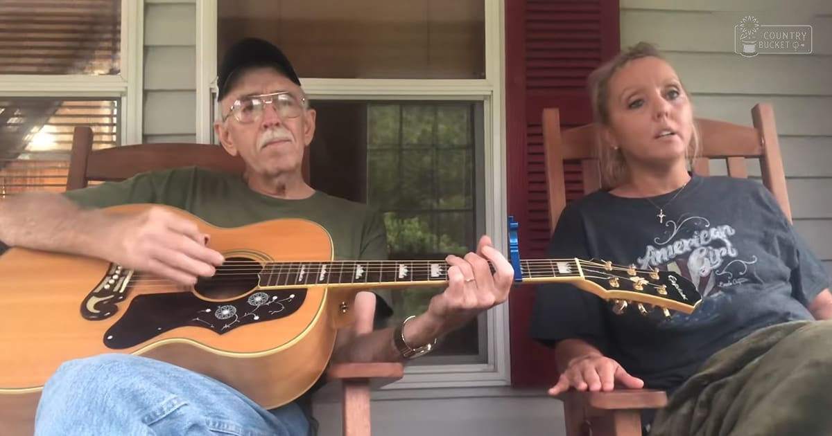 An older adult male singer playing a guitar with an adult female sitting beside him in a porch setting, wearing casual attire and accessories.