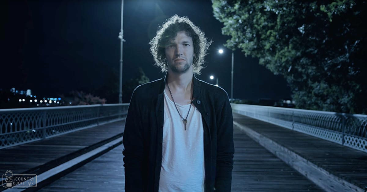 A young adult male wearing a dark jacket over a white t-shirt with an ornate metal bridge railing and streetlights in the background at night.