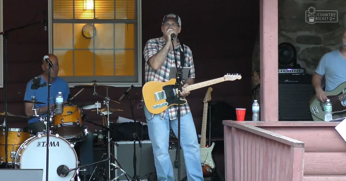 Middle-aged man playing guitar and singing into a microphone at an outdoor concert with other musicians, rustic backdrop, daytime lighting.