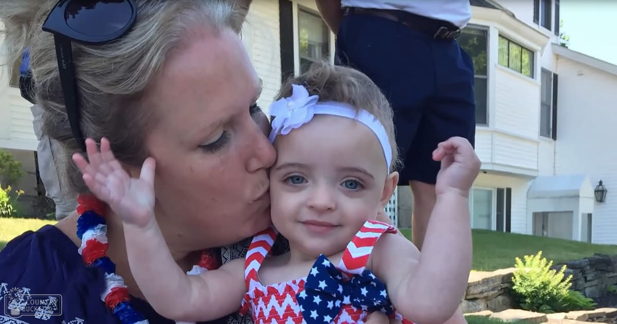 A middle-aged woman holding a toddler in front of a suburban home, wearing dark blue with white patterns, red sunglasses, and streamers; the toddler is in a red and white chevron-patterned dress with star patterns and a white headband.