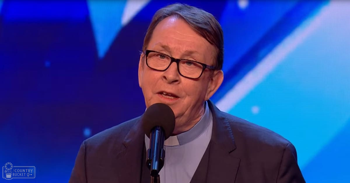 Middle-aged male in a studio setting, wearing a dark suit and light collared shirt with a clerical collar. Microphone and geometric screen backdrop suggest a professional broadcast environment.