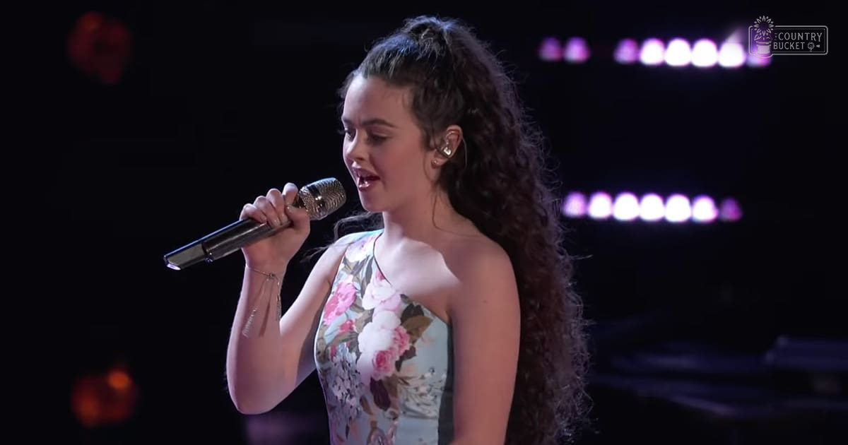 A young adult female singer wearing a strapless floral dress performs on stage under dim lighting.