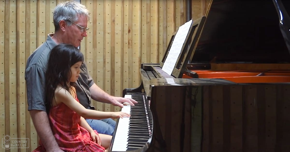 A middle-aged man teaching a toddler to play piano in a music studio with wooden paneling and sheet music on an open stand above the grand piano.