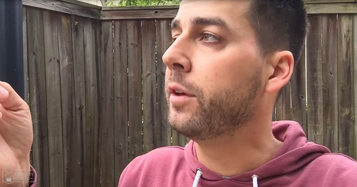 A young adult male wearing a maroon hoodie stands outdoors in spring or summer, with soft natural light and a wooden fence backdrop.