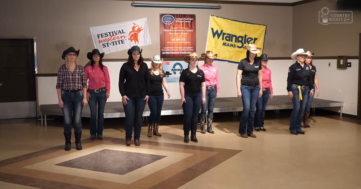 Group photo at Festival Western St-Tite event indoors with neutral walls and stage-like platform. People wearing cowboy boots and casual attire in a community center setting.