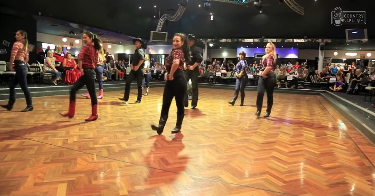 A dance group performing on a wooden ballroom floor with an audience seated around them, under overhead lights.