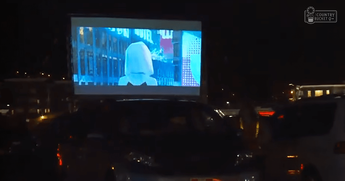 An outdoor drive-in movie theater at night, with a film screen displaying 'Madly Odd', surrounded by parked cars and other vehicles under artificial lighting.