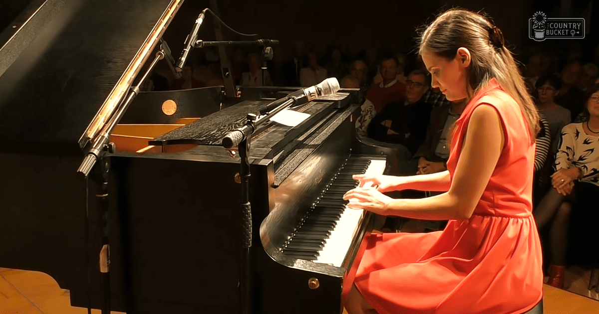 A young adult female plays a grand piano in an indoor concert setting with dim evening lighting.