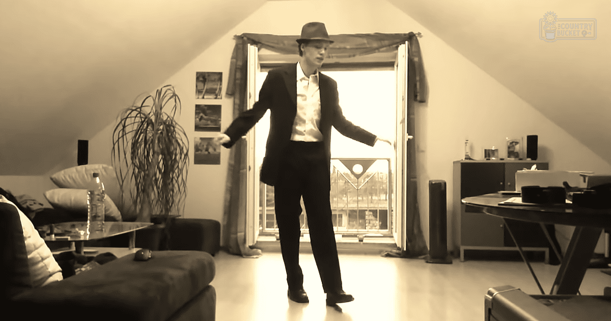 Young adult male wearing a dark suit jacket and fedora hat, seated amidst attic-like furniture with sloped ceiling and natural light.