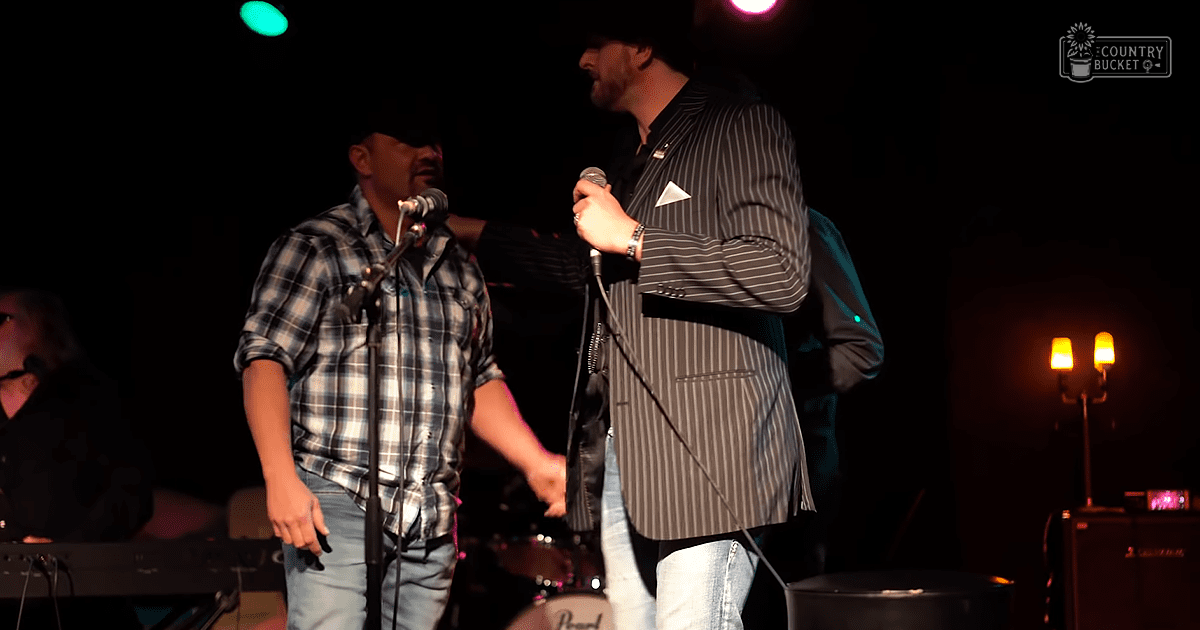A middle-aged to older adult male singer in a striped blazer and jeans performs at an indoor venue, holding a microphone; another adult male in a plaid shirt and jeans stands nearby. The setting includes microphones, musical instruments, amplifiers, and stage lights with colored beams.