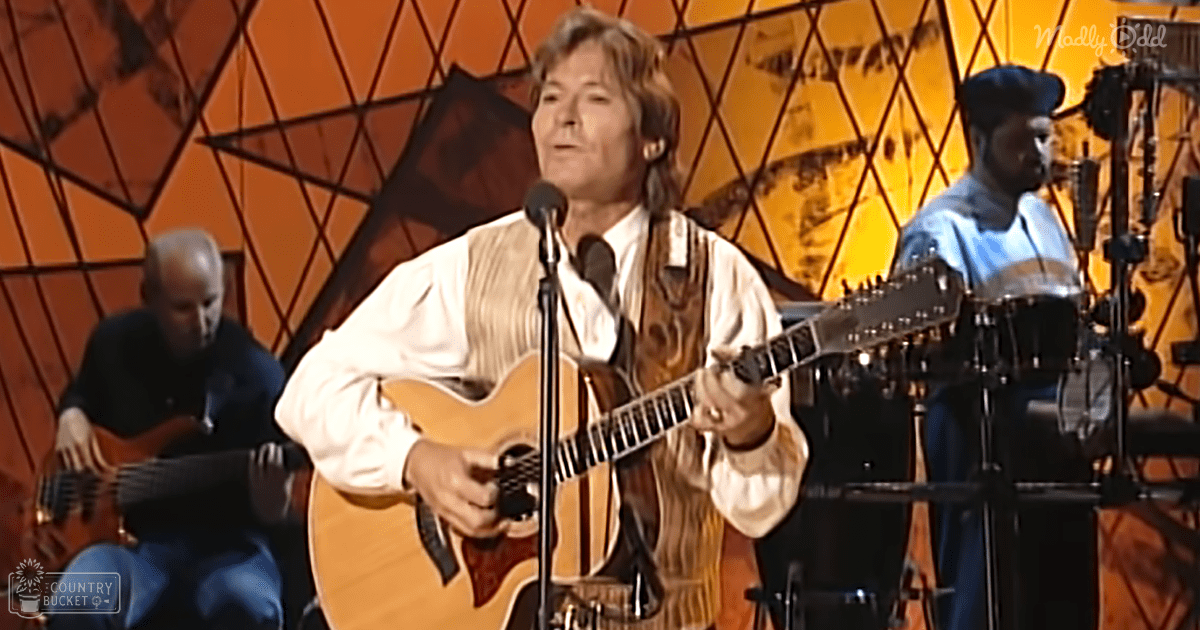A middle-aged man wearing a white shirt and textured vest plays guitar while seated next to an older adult male, both on stage at what appears to be a music event with microphones and drums in the background.