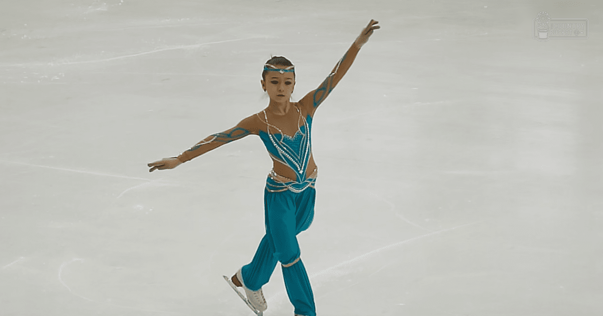 A young female figure skater in a teal costume with intricate patterns and embellishments stands on an indoor ice rink, wearing white ice skates and a matching headband.