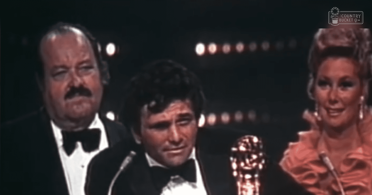A man in a black tuxedo holding an Oscar statue at the Academy Awards ceremony, with stage lighting and other attendees in formal attire.