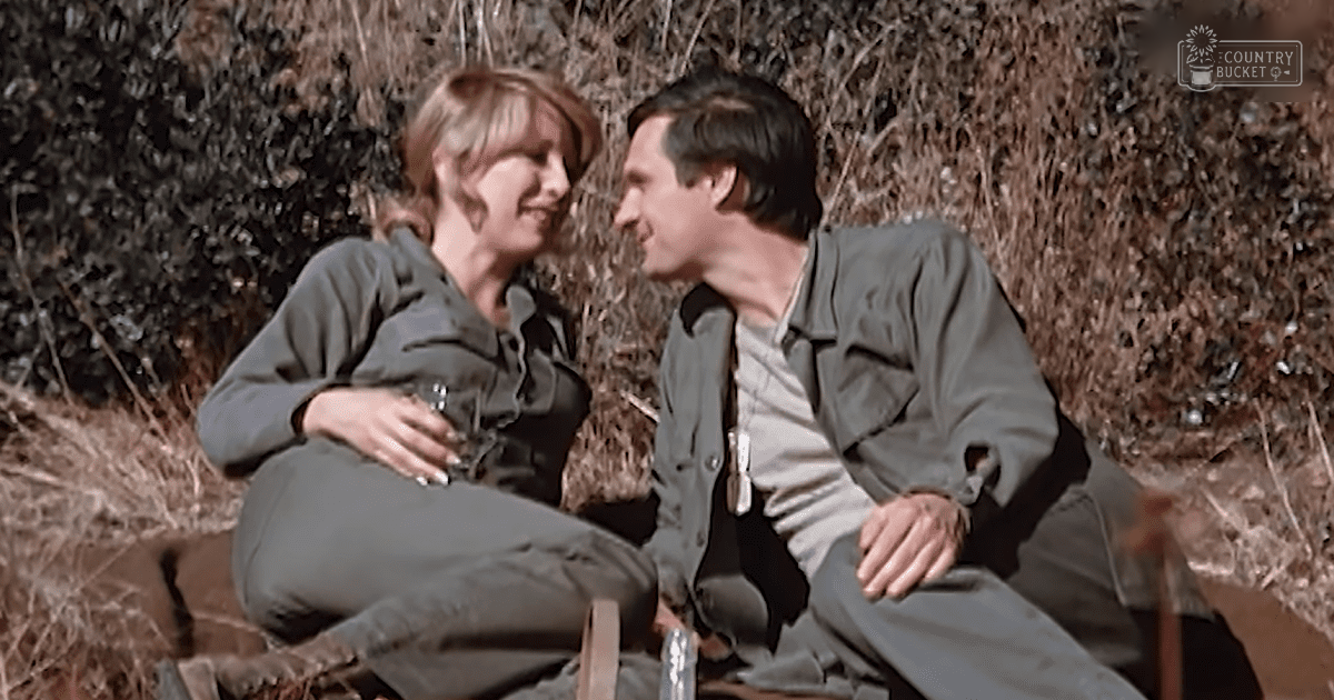 A couple outdoors with autumnal foliage and outdoor furniture, wearing casual attire under natural daylight.
