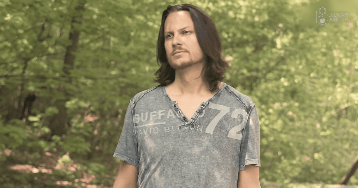 A young adult male stands outdoors amidst a forested area with green foliage, wearing a modern casual grayish-blue v-neck t-shirt.