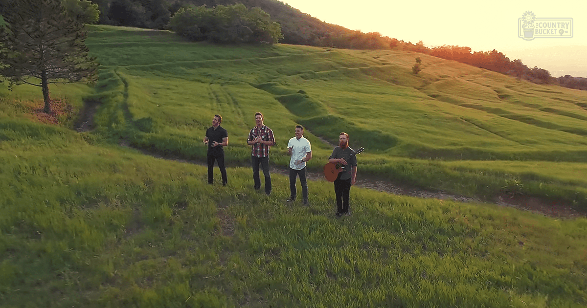 Four young adult males singing in an open field during sunset with green grass and rolling hills.