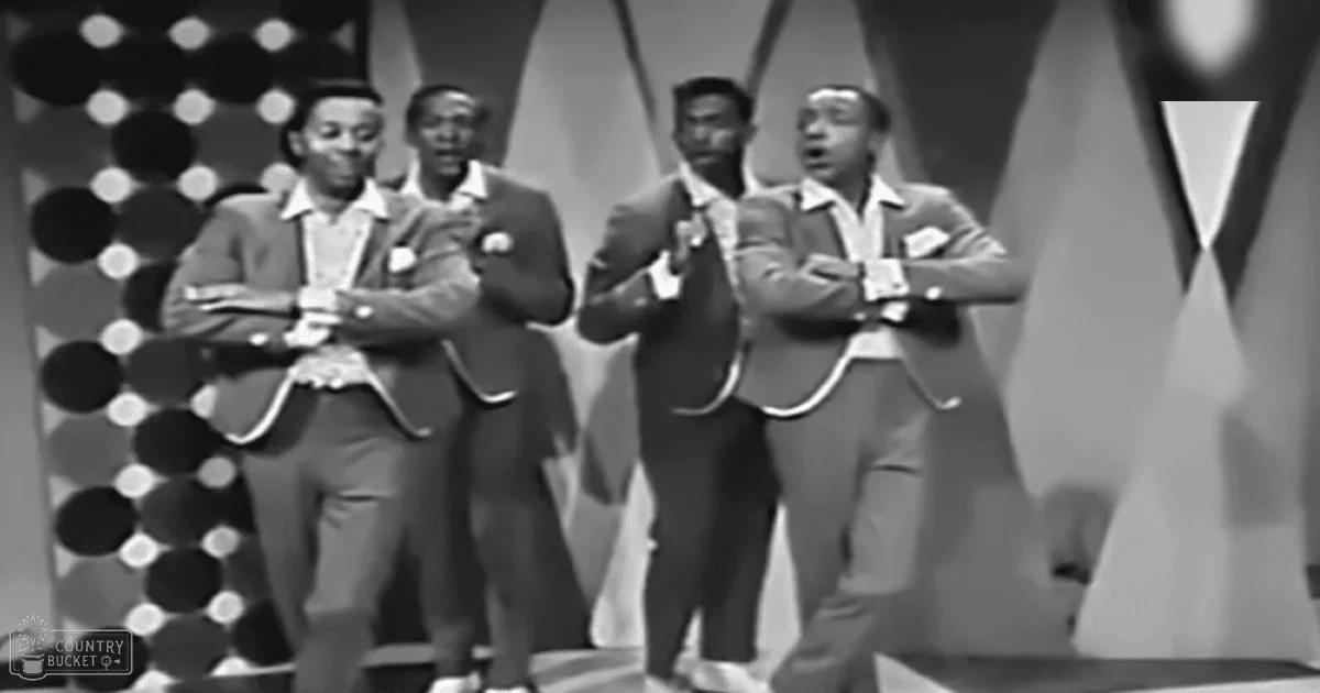 Adult males in suits performing on a stage with geometric-patterned backdrop indoors under artificial lighting.