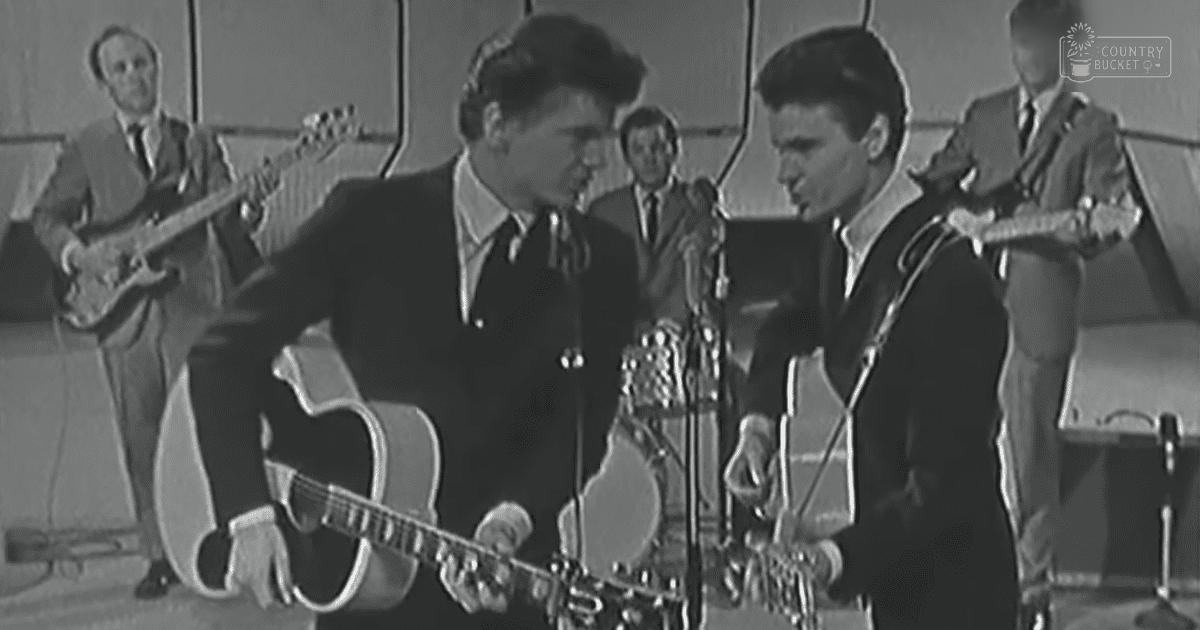 A young adult male plays guitar on a stage with another person nearby holding an instrument; background is simple, possibly indicating a live music performance.