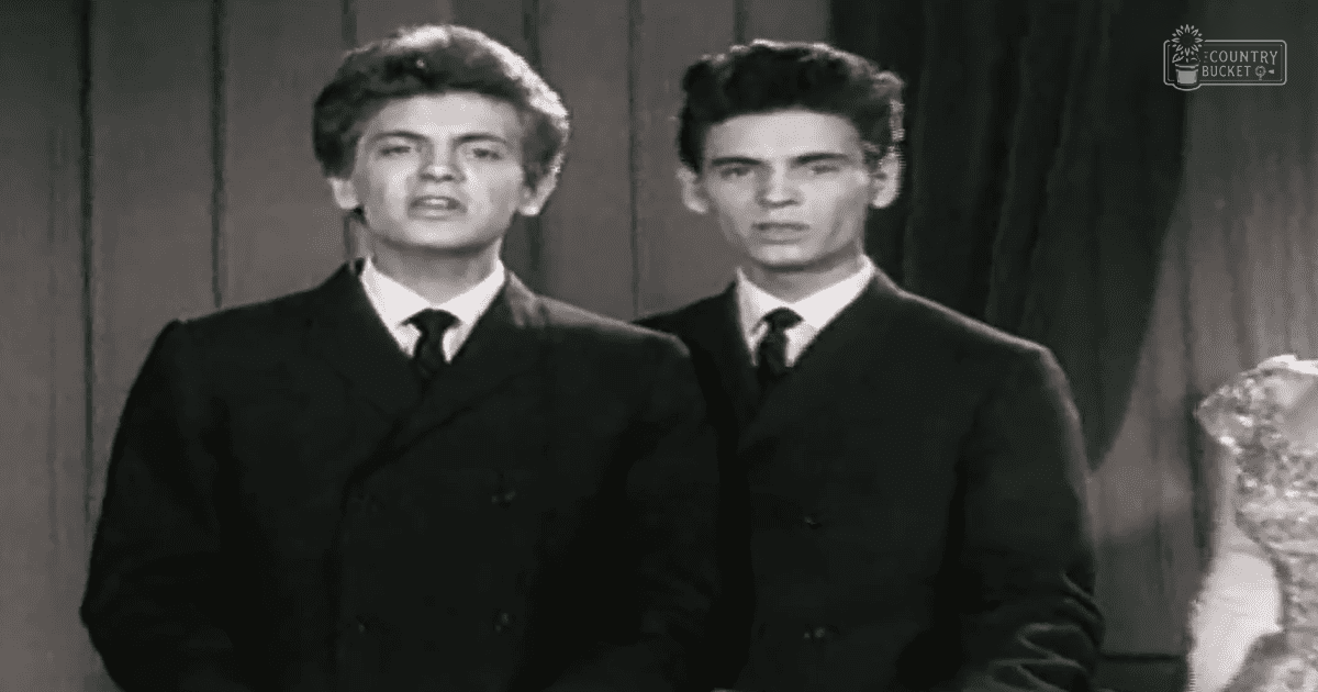 Two young adult males in a studio setting, dressed in dark double-breasted suits with white dress shirts and black ties, surrounded by vertical wooden panels or curtains.