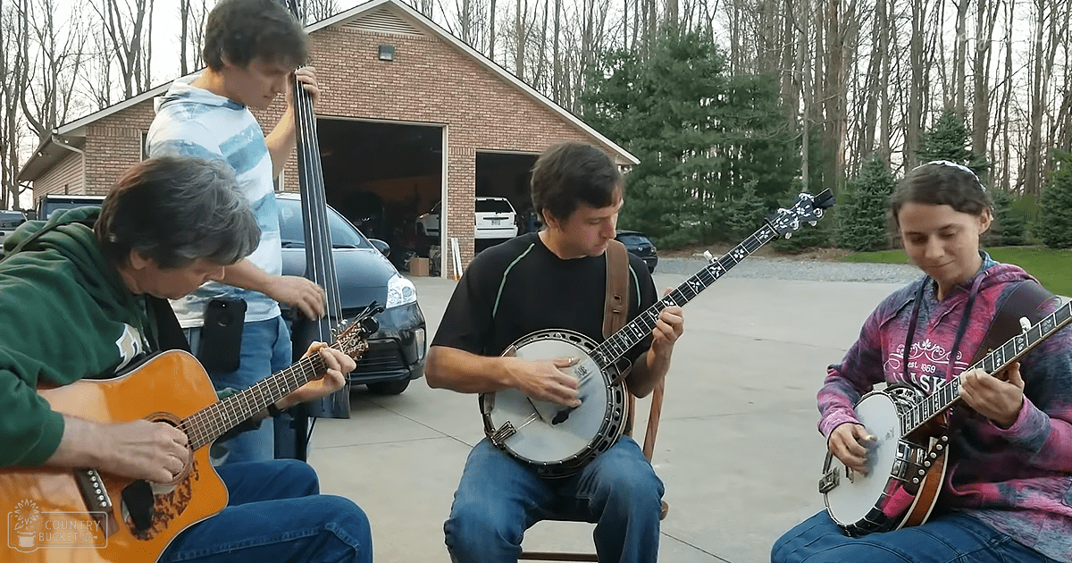 Four teenagers playing musical instruments on a residential driveway during late autumn or winter, with brick house and parked cars visible.