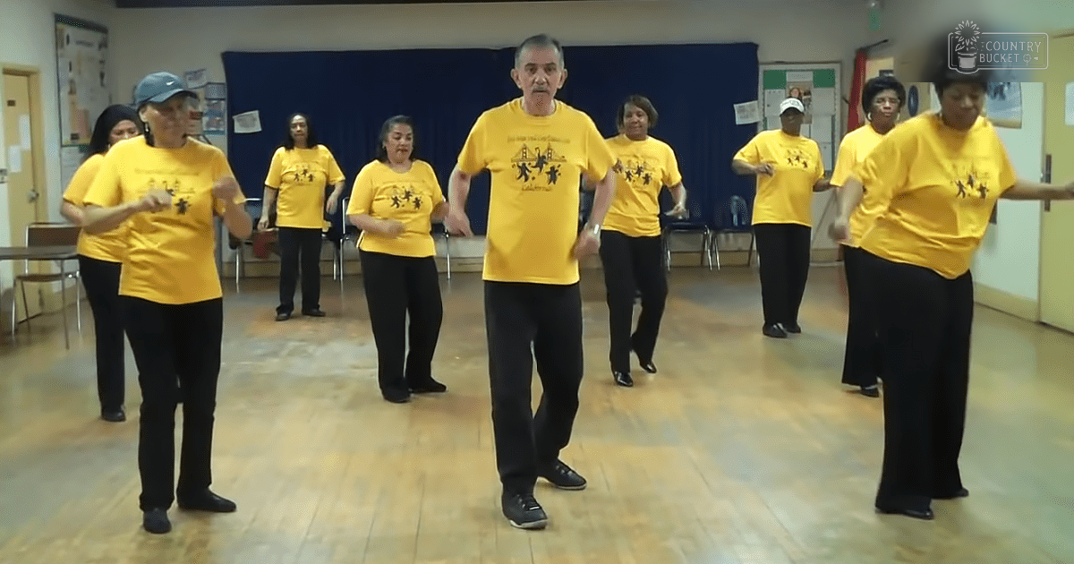 A group of middle-aged to older adults wearing matching yellow T-shirts with a graphic design, gathered in an indoor space possibly a community center or dance studio. The setting includes wooden floors and chairs against the wall under blue curtains.