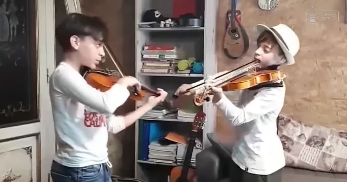 Two young male violinists in a casual home setting, one wearing 'CAPTAIN' shirt and jeans, the other in white shirt and dark pants with hat; bookshelf, guitar, cushion with patterned design visible.