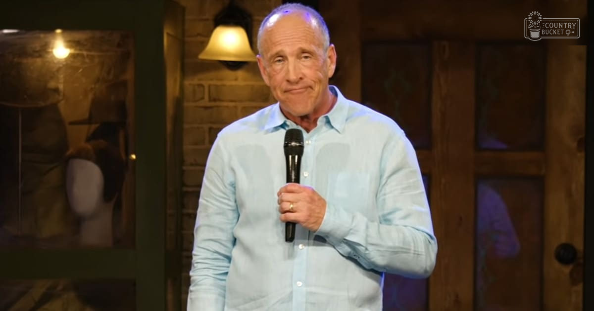 A middle-aged adult male in a relaxed fit, light blue button-up shirt with a collar, holding a microphone indoors at what appears to be a comedy club or theater setting.