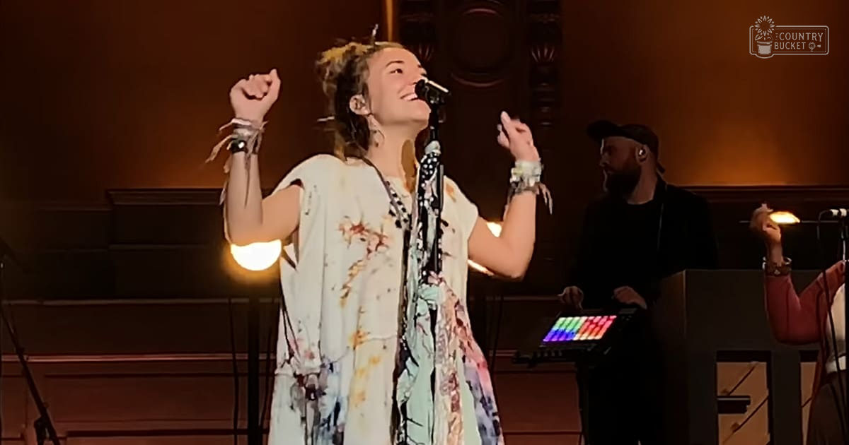 A young adult female performer wearing a loose, tie-dye dress and multiple bracelets on her wrists, performing in an indoor venue with warm lighting.