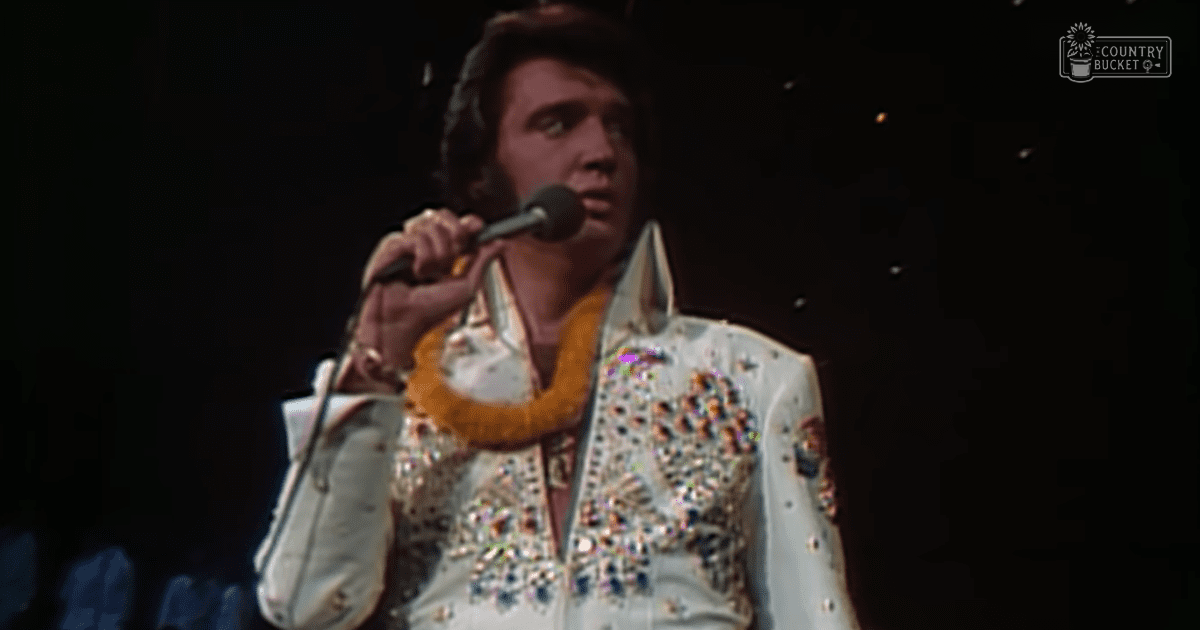 A male adult singer in a white, sequined 1970s-style stage jacket with numerous shiny embellishments performs live at an indoor concert venue under stage lighting.