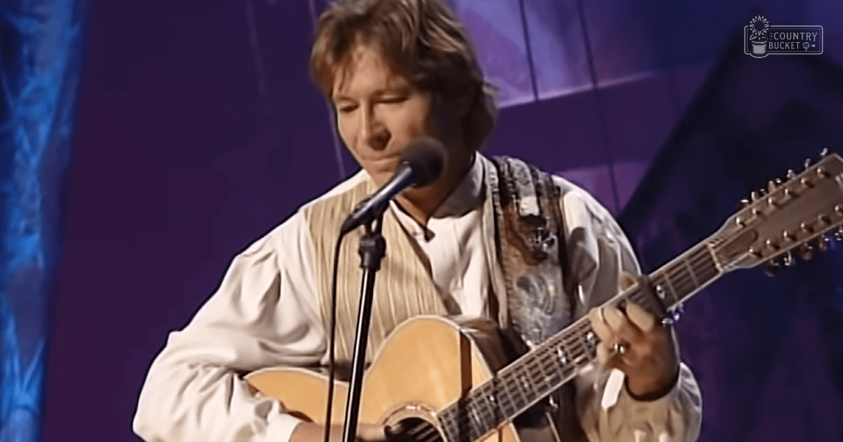 A young adult male singer playing an acoustic guitar, wearing a beige long-sleeved button-up shirt with vertical stripes and holding a light brown classical guitar in front of a microphone on stage.