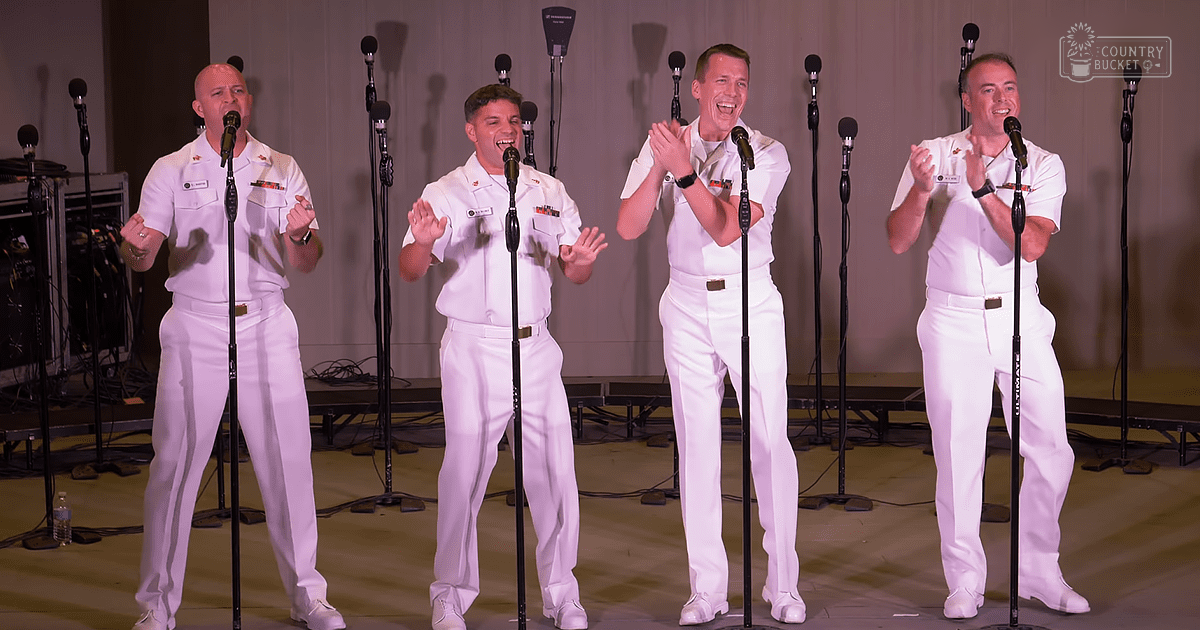 Group of adult males in white naval uniforms performing on a stage with microphones, in an indoor venue with artificial lighting.