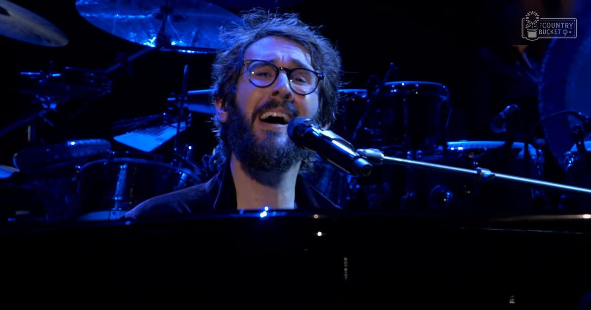 Adult male singer performing at a concert venue with dark attire and stage lighting, blue piano keyboard in view.