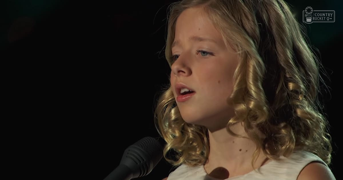 A young female child singing into a microphone indoors, wearing a white sleeveless dress with dimly lit background.