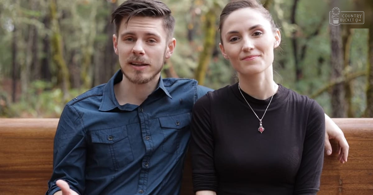 A young adult couple in a park setting with green foliage, wearing casual attire. The man wears a dark blue button-up shirt, while the woman is dressed in black and accessorized with earrings and a red gemstone pendant.