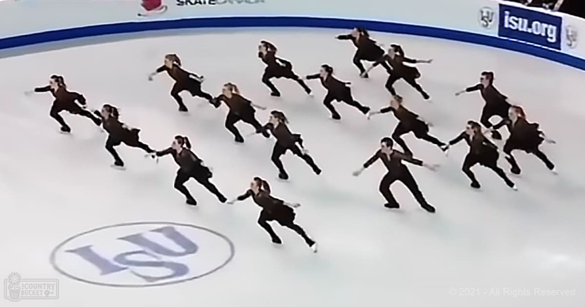 Synchronized skating team performing at an indoor ice rink with markings and ISU logo signage, well-lit setting.