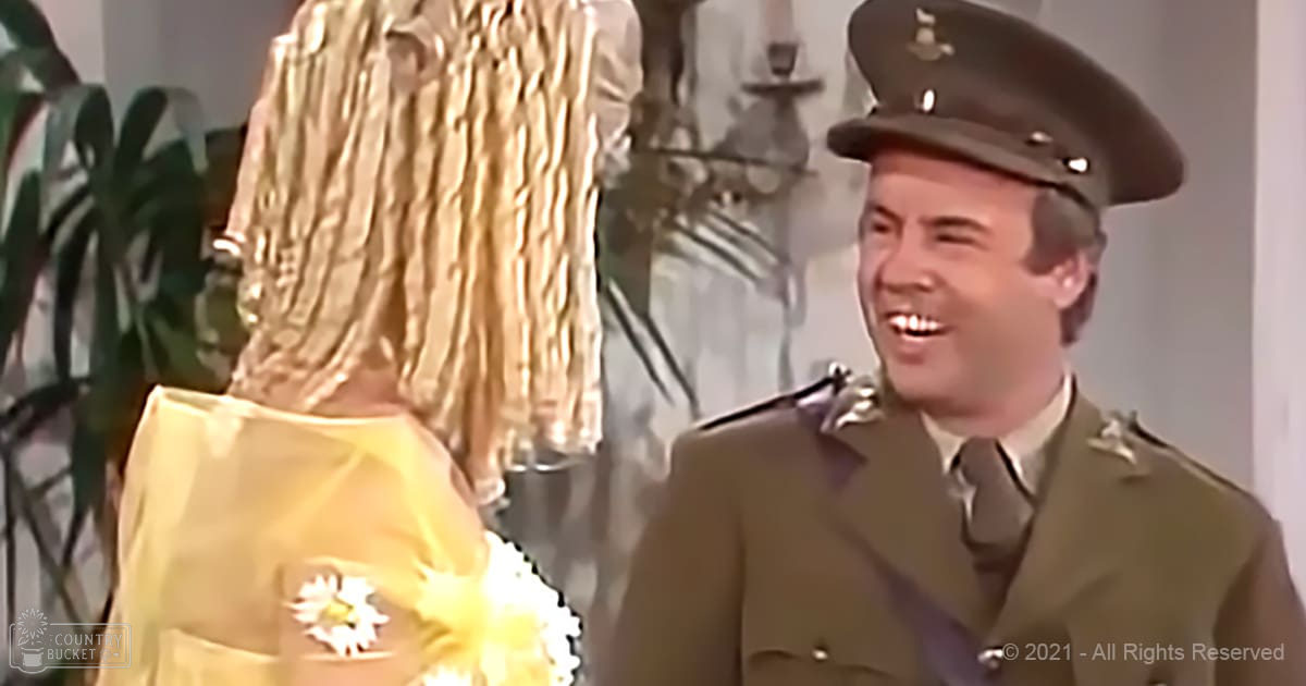A man in a mid-20th-century military uniform stands indoors with an olive green jacket and peaked cap adorned with insignia, surrounded by neutral tones and subtle decorative elements.