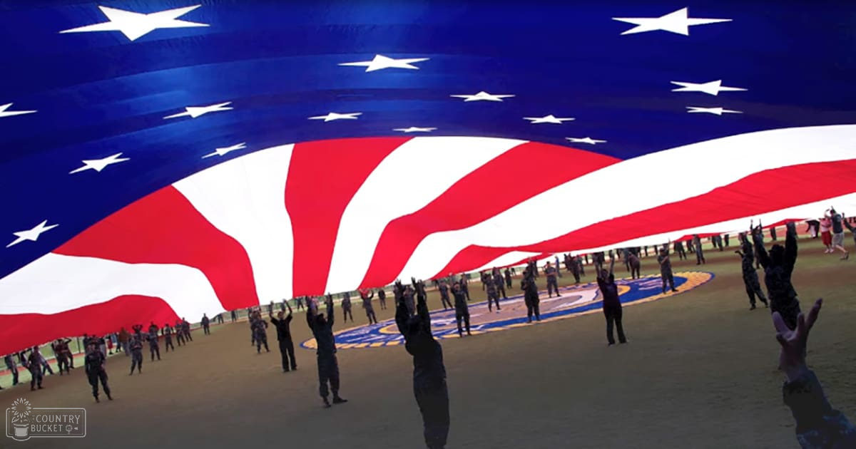 Military personnel holding a large American flag at an airfield during what appears to be a ceremonial event, with uniformed young adults and blue skies overhead.