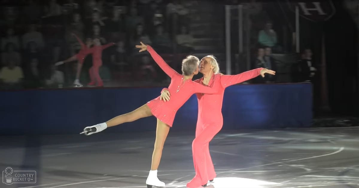 A middle-aged to older adult female and male figure skating duo performing at an indoor ice rink with red outfits, white figure skate boots, and wrist guards under artificial lighting.