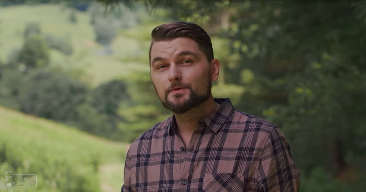 Adult male in a plaid button-up shirt outdoors with greenery and hills in the background during daytime.