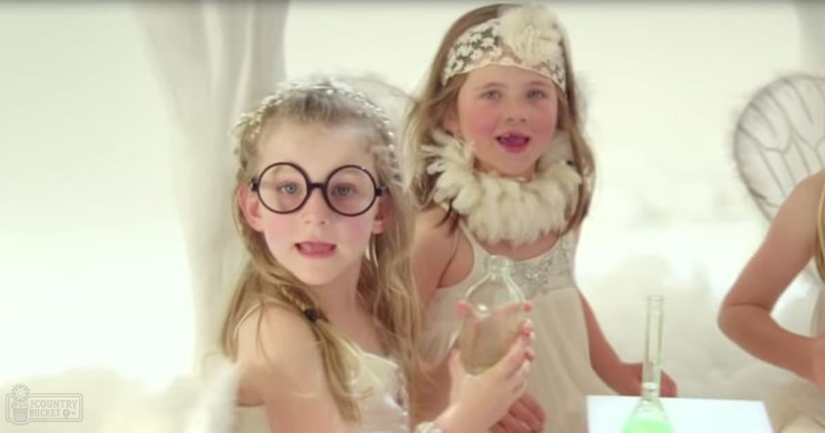 Two young children, one wearing a white lace dress with large round glasses, the other in a light-colored dress with fur-like collar and headband, stand indoors under bright daylight.
