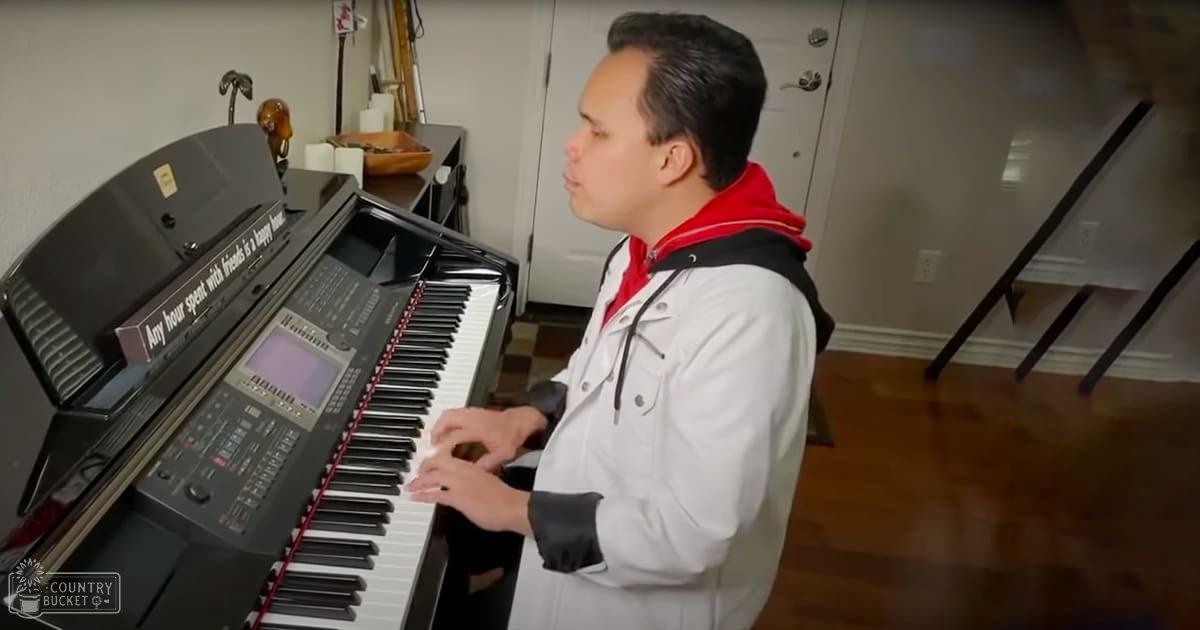 A young adult male in a relaxed modern casual wear jacket with black and red accents plays a digital keyboard indoors, surrounded by home decor elements.