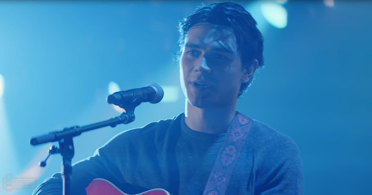 A young adult male wearing a relaxed dark gray or blue long-sleeved shirt with no visible pattern, performing in an indoor stage setting under blue lighting.